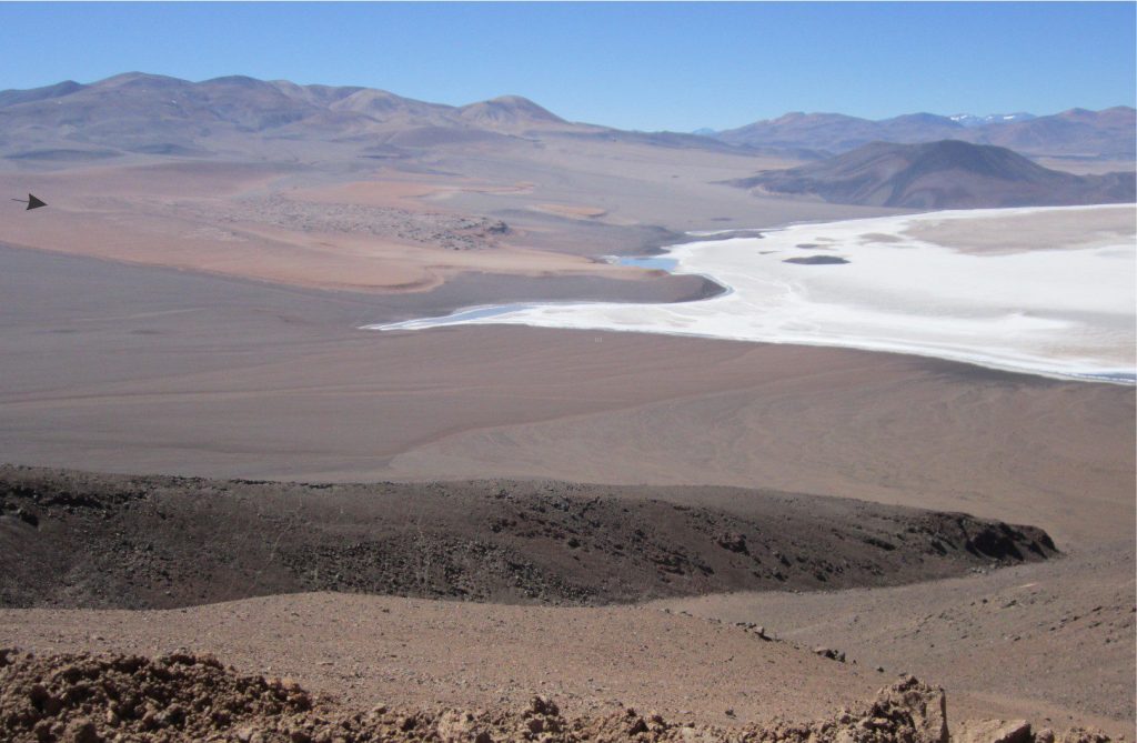 Eroded remnants of a Middle Miocene welded ignimbrite bordering Salar LaGrande, Chilean Altiplano (orange hues, arrow top left). The high ground behind the ignimbrite is mostly composite andesitic and basaltic cones of similar age. The eruptive episodes were associated with Mid-Miocene caldera collapse. The ignimbrite is lapped by more recent, arid, alluvial fans, the alluvium derived from Mid Miocene basalts and andesites. The Salar is capped by thick (white) gypsum and halite crusts.