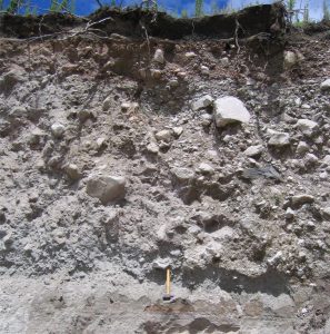 A block and ash flow derived from dome collapse at Mt. Tarawera. Note the angularity of blocks and coarse lapilli (to 50 cm wide in this view), extremely poor sorting, and lack of preferred clast alignment. The stratified ash beneath the BAF may be a surge deposit. Hammer lower centre. Image credit: Tarawera, Kari Cooper, 2017, National Science Foundation, public domain 