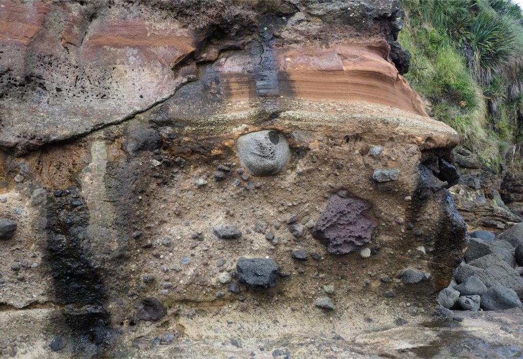 Andesite boulders up to 40 cm across protrude through the upper bedding plane of a lahar where they are draped by later, airfall ash and lapilli (white to red-brown layered beds near the top of the exposure). Pliocene Karioi volcano, Raglan, New Zealand.