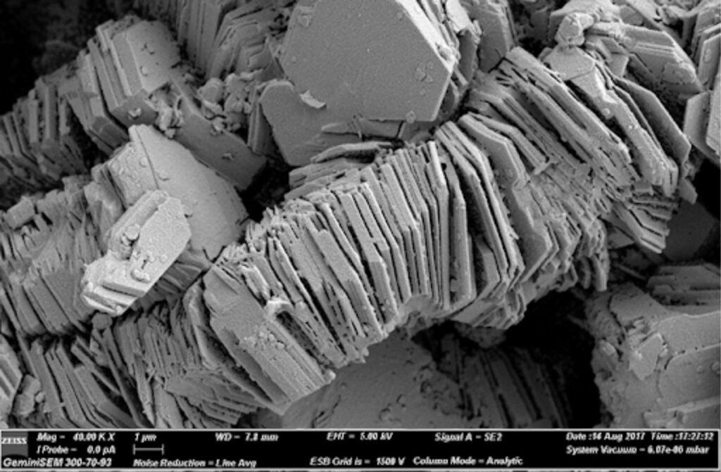 An SEM image of kaolinite crystals stacked along the c-axis into book-like structures – a common crystal habit for this mineral. Bar scale 1μm is on lower left.  Image credit: Open Geology: Earth Materials – The Rock Forming Minerals, CC-BY.